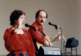 A woman in a dress speaks into microphone on stand with her hand open near face. Man to the rig…