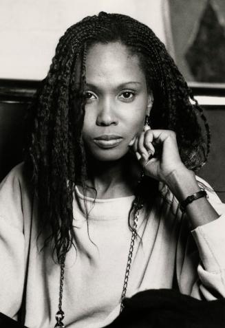 young woman with braided hair and chain necklaces in white sweater has hand to face. 