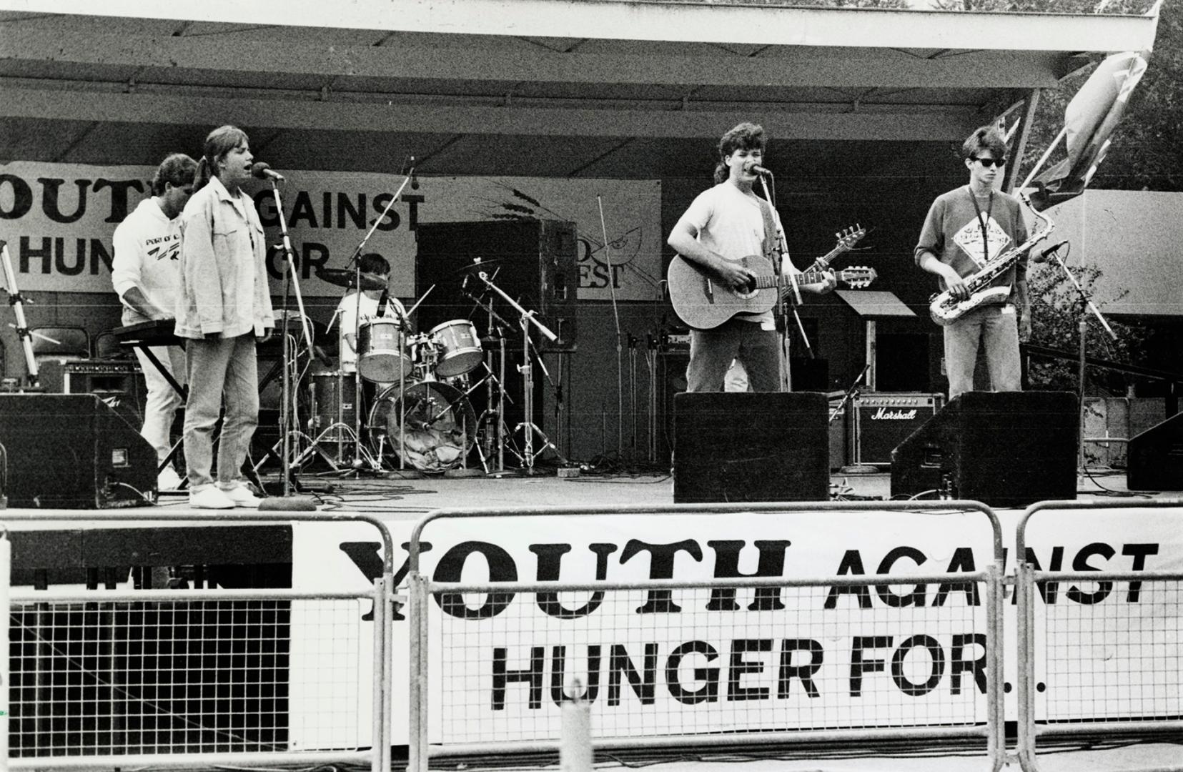 A band performing on stage. There is a banner stating Youth against hunger for...