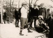 A group of people stand and sit outdoors in the snow, dressed in winter clothes. The central fi…