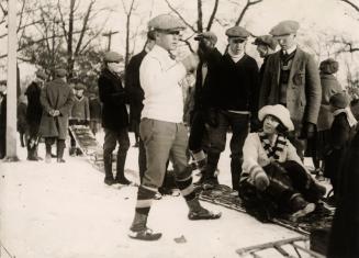 A group of people stand and sit outdoors in the snow, dressed in winter clothes. The central fi…