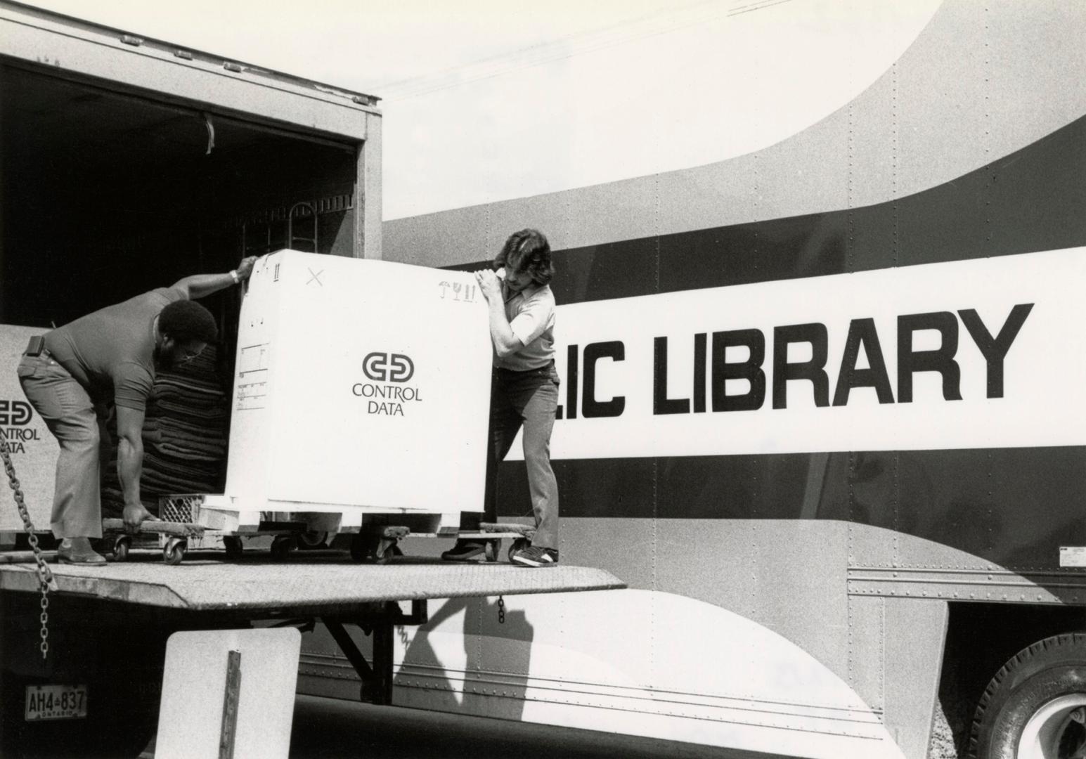 Picture of two men unloading a large box off the back of a truck. 
