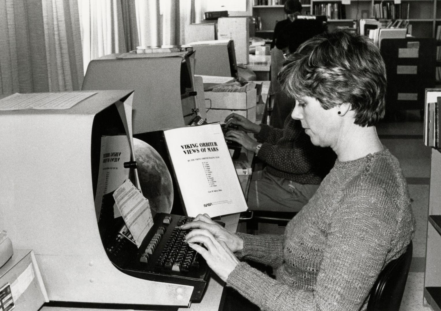 Picture of a woman typing on a keyboard at a large computer terminal. 