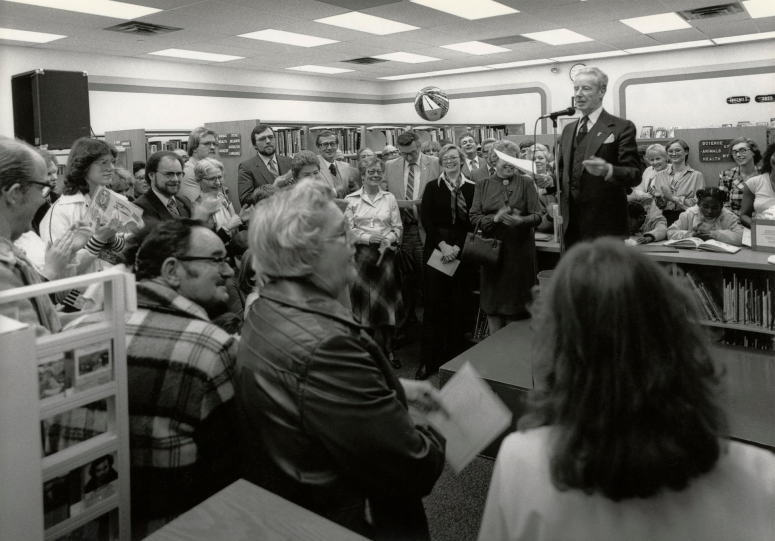 Picture of man talking at a microphone in front of crowd of people in a library. 