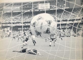 A soccer ball hits the back of the net. Behind it the goalie lies on the grass and other player…