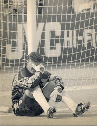 Man in soccer shoes, short and jacket sits in the soccer net on the ground with his gloved hand…