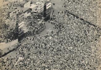From Above, thousand of people throng in square with podium on left. 