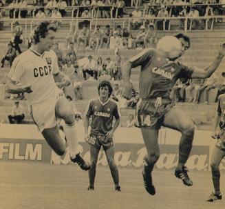 Soccer stadium. On the field CCCP player legs twisted in the air, Canadqa player in the air bal…