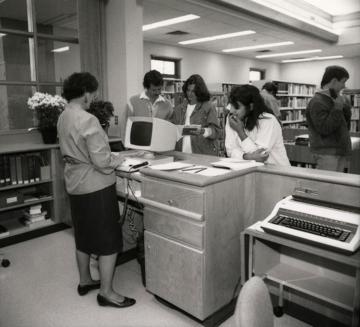 Picture of a library staff member standing at a desk with a computer and three customers in fro…
