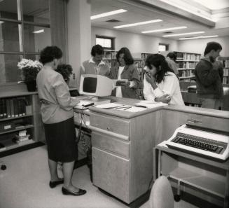 Picture of a library staff member standing at a desk with a computer and three customers in fro…