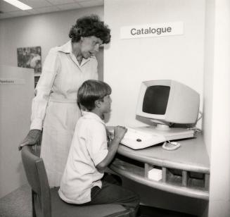Picture of a library staff member standing standing at a computer catalogue with a young boy se…