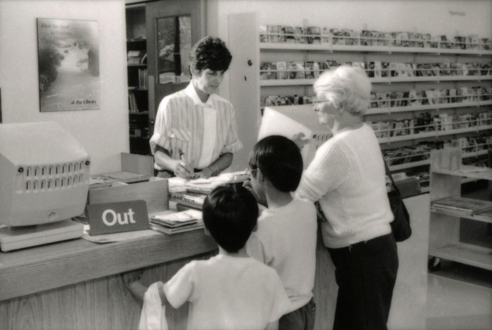 Picture of a library staff member standing at a desk checking out books for a woman and two chi…