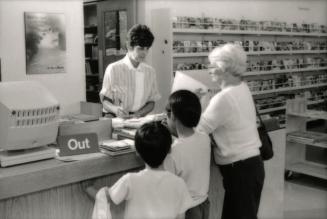 Picture of a library staff member standing at a desk checking out books for a woman and two chi…