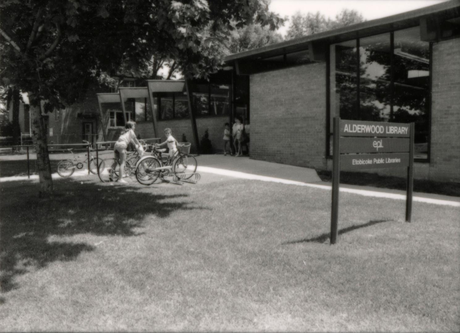 Picture of exterior of one story library with wide concrete path. Bicycles and people are visib…