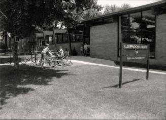 Picture of exterior of one story library with wide concrete path. Bicycles and people are visib…
