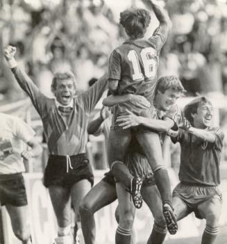 Soccer players: from left one runs in with fists in the air, player lifts another off the groun…