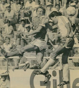 In front of audience in stands two soccer players jump in the air for the same ball. 