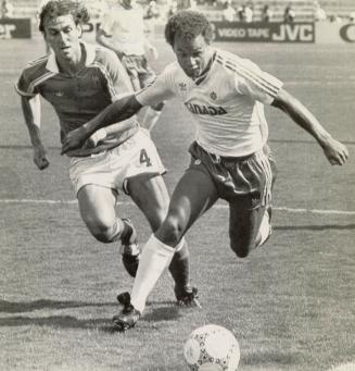 Soccer Player in Canada jersey hurtles down the field with the ball in front of him pursued by …