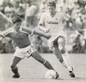 Soccer players on field. French player on left has ball on his toe while Canadian runs up to th…