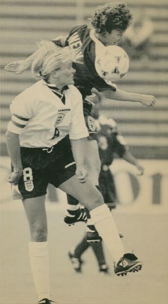 Woman on left with one foot up looks toward right at another woman in the air with the ball at …