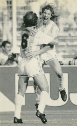 On the soccer field, two men in soccer gear. Man leaps into the arms of teammate. His mouth is …