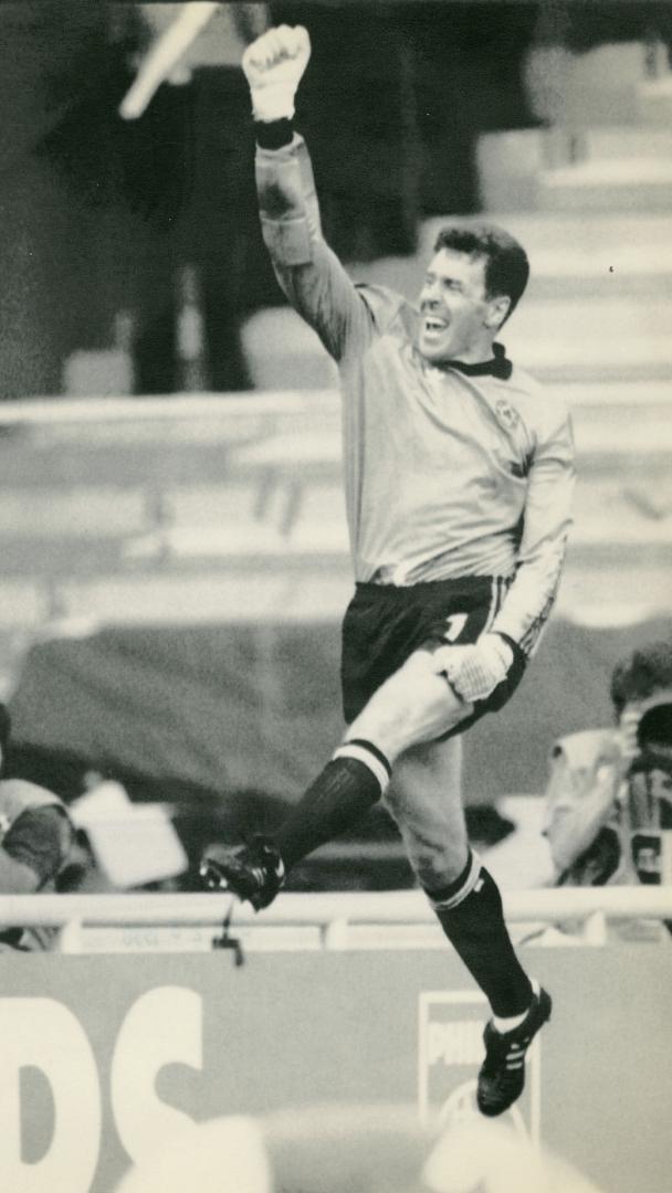 Full body shot, man in soccer gear leaping with legs in scissor kick and arm and fist hoisted a…