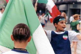 Teen male on left has the word Italy shaved onto his head. He stands in front of an Italian fla…