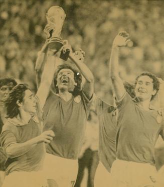 Three men in soccer gear in front of a large audience. The middle one holds up the World Cup tr…