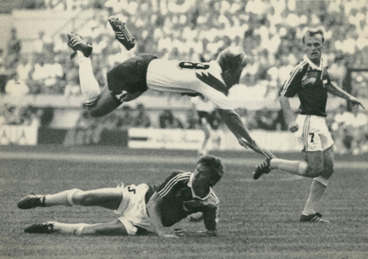 On soccer field, three men in soccer gear. One flies over another who is on his side on the gro…