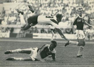 On soccer field, three men in soccer gear. One flies over another who is on his side on the gro…