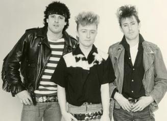 Three young men in jeans with haircuts, shaved on side and spiky or curly on top. Middle is blo…
