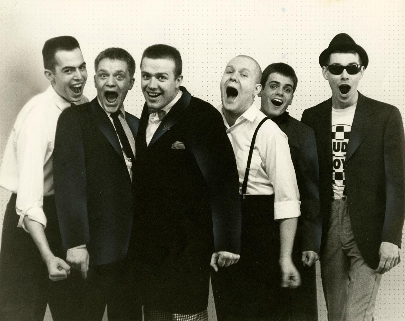 six young men with mouths open as if singing. Against a white background, they wear dark trouse…