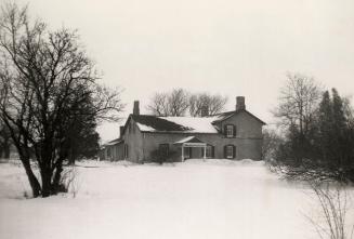 Picture of a two storey farmhouse with field of snow and trees in foreground. 