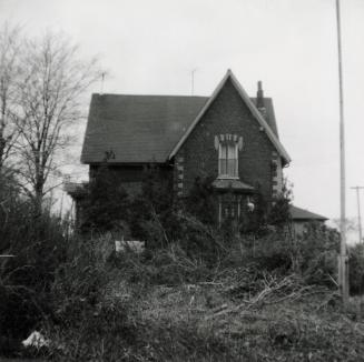 Picture of a brick farmhouse with peaked roof and vegetation in front. 