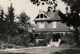 Picture of a two storey house with large veranda, peaked roof and children sitting out front an…