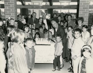 Picture of a crowd of people gathered around a large cake and a woman cutting into the cake wit…