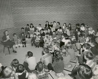 Picture of a librarian reading to a group of children seated in a circle around her. 
