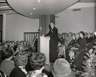 Picture of a man speaking at a podium and a crowd of people seated behind and in front of him. 
