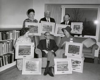 Picture of a man seated in a chair surrounded by five people kneeling and standing holding fram…