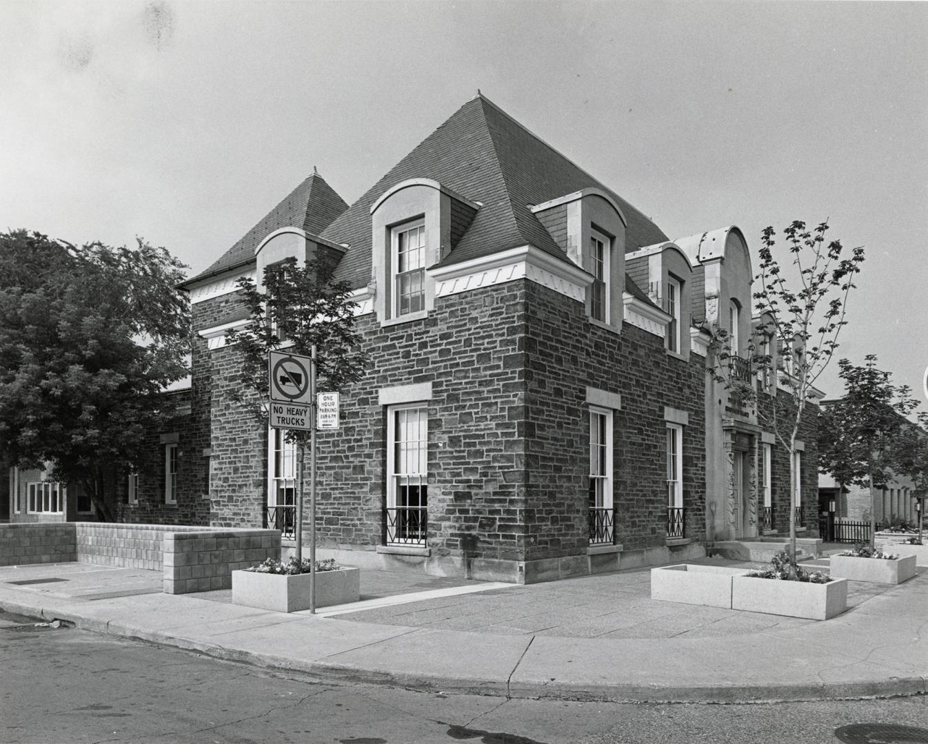 Side corner view of two storey stone library building with trees in planter boxes in front and …