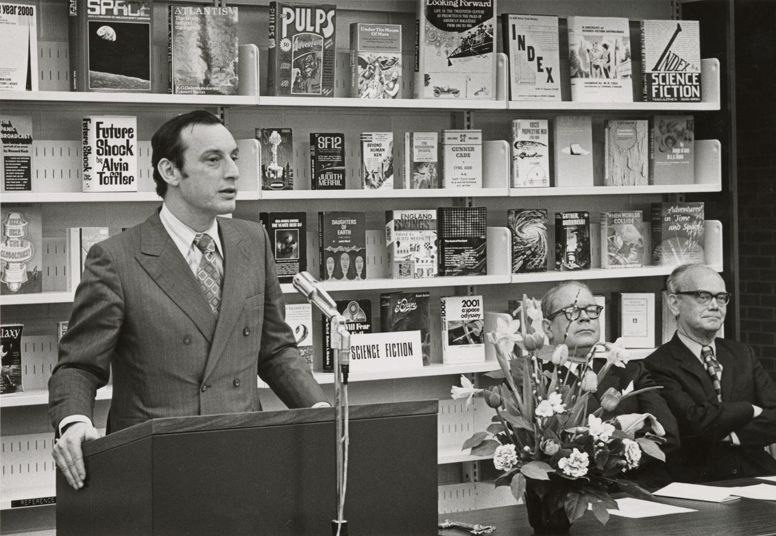 Picture of a man standing speaking at a podium with a bookshelf behind him and two men seated a…