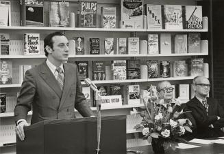 Picture of a man standing speaking at a podium with a bookshelf behind him and two men seated a…