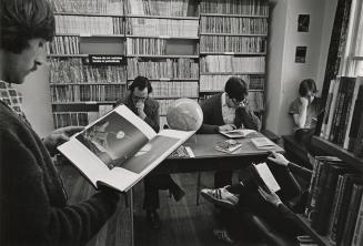 Picture of a man standing reading a book with others seated reading at a table in a library. 