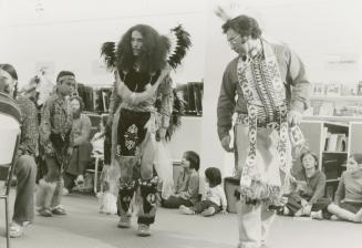 Picture of three Indigenous dancers with children sitting on the floor watching. 