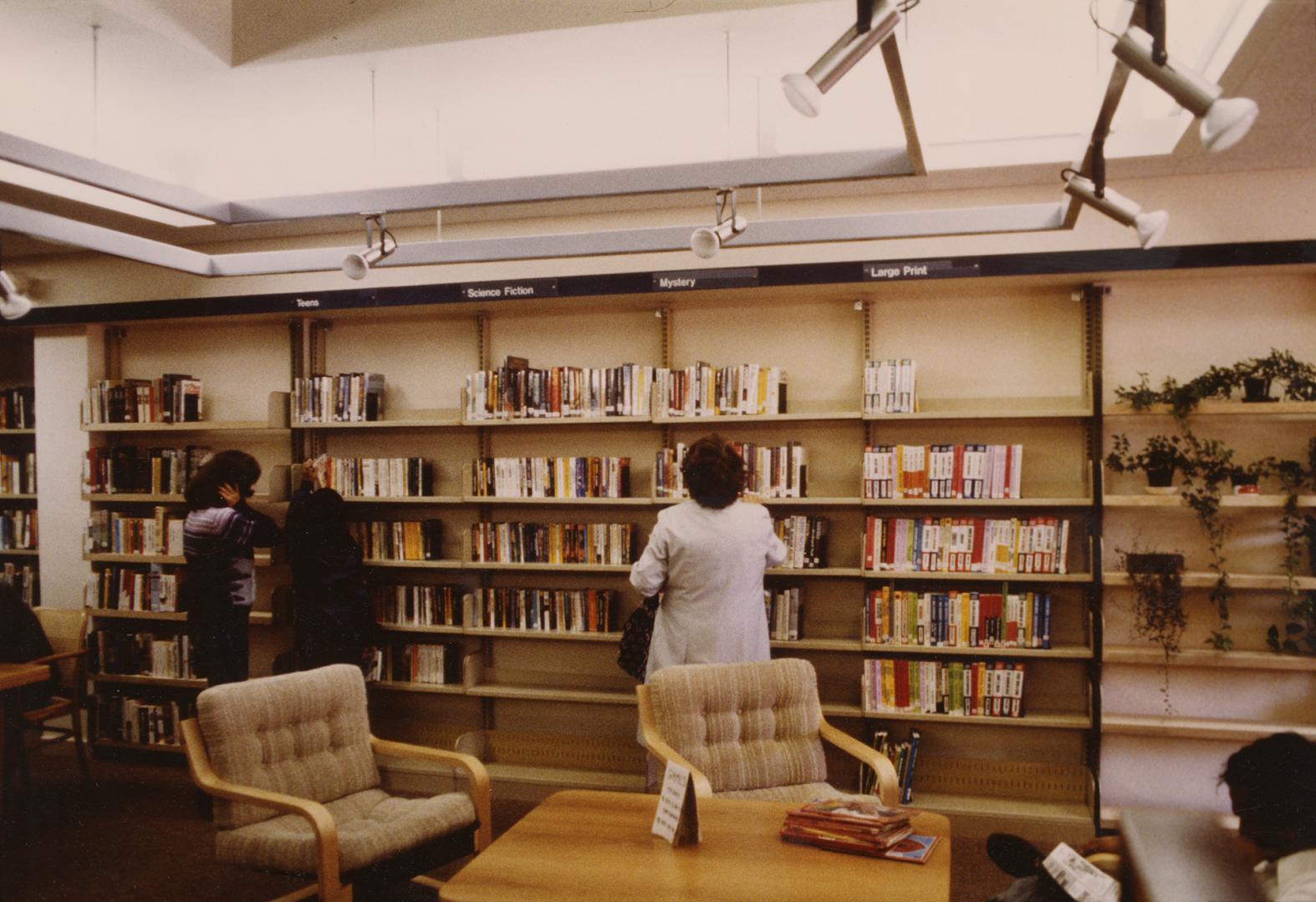 Picture of library interior showing people browsing shelves of books and table and two chairs i…
