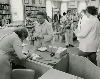 Picture of a room in a library with a librarian writing at a desk and older woman standing in f…