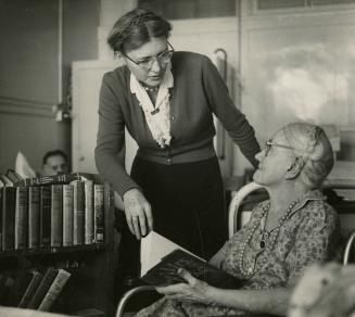 Picture of a librarian with a book truck leaning over an older woman who is seated with a book …