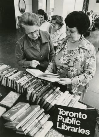 Picture of a librarian and another woman stand look at a book from a full book truck. 