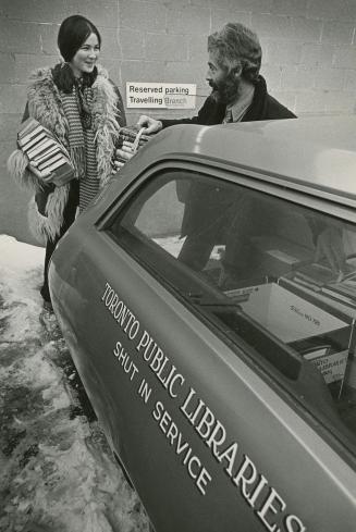 Picture of a woman holding piles of books and a man helping put them in the back of a car that …