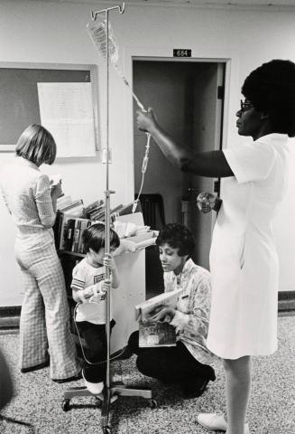 Picture of a librarian crouching on a hospital floor showing a book to a small boy who is hooke…